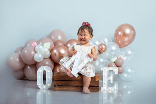 A joyful baby girl sitting on a wooden crate with decorative balloons and the word ONE. Where to Buy 1st Birthday Helium Balloons?